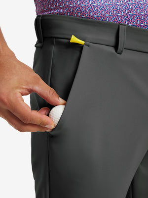 Hand placing a golf ball into pocket of water-resistant golf stretch nylon pants in dark grey.